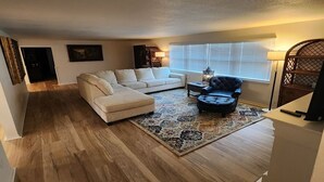 A living room with a sectional sofa, a black leather armchair, a patterned rug, and a window with blinds.