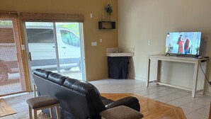 A living room with a black leather sofa, wooden floor, a television, and a window with a view of the street. - Wonderful 3 bedroom house (Scott)