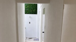 Un couloir étroit aux murs blancs et au sol carrelé, qui mène à une porte. On aperçoit un mur vert à travers la porte entrouverte.