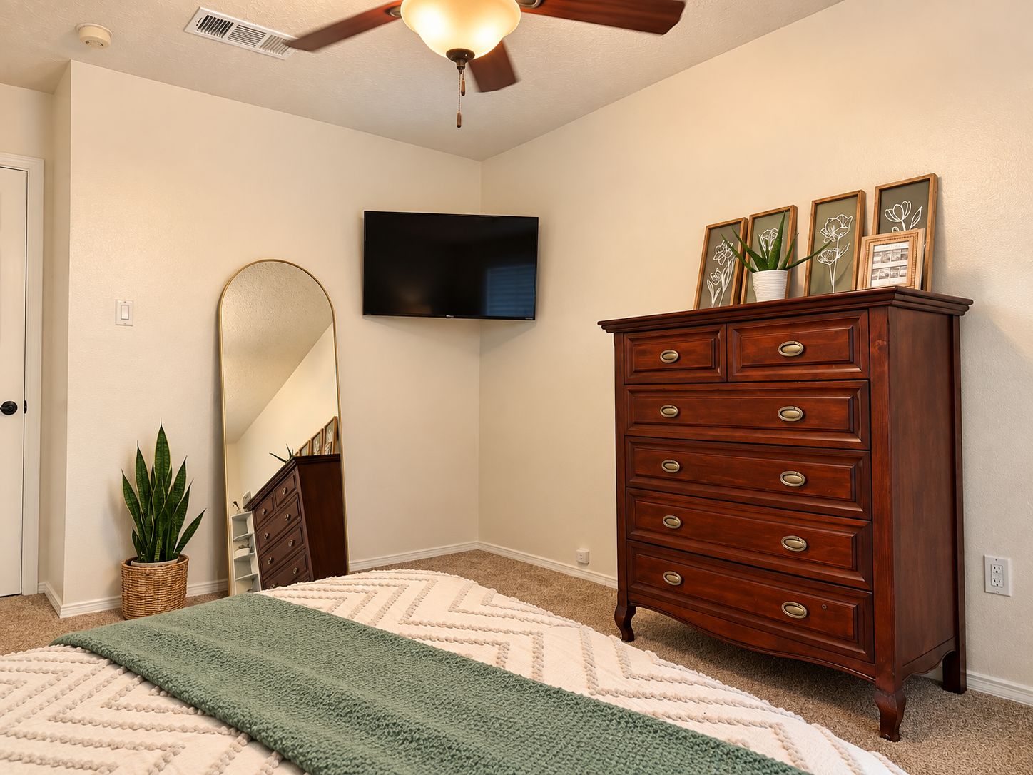 A bedroom with a ceiling fan, a flat-screen TV mounted on the wall, a wooden dresser with drawers, a mirror, a green and white rug, and a potted plant.