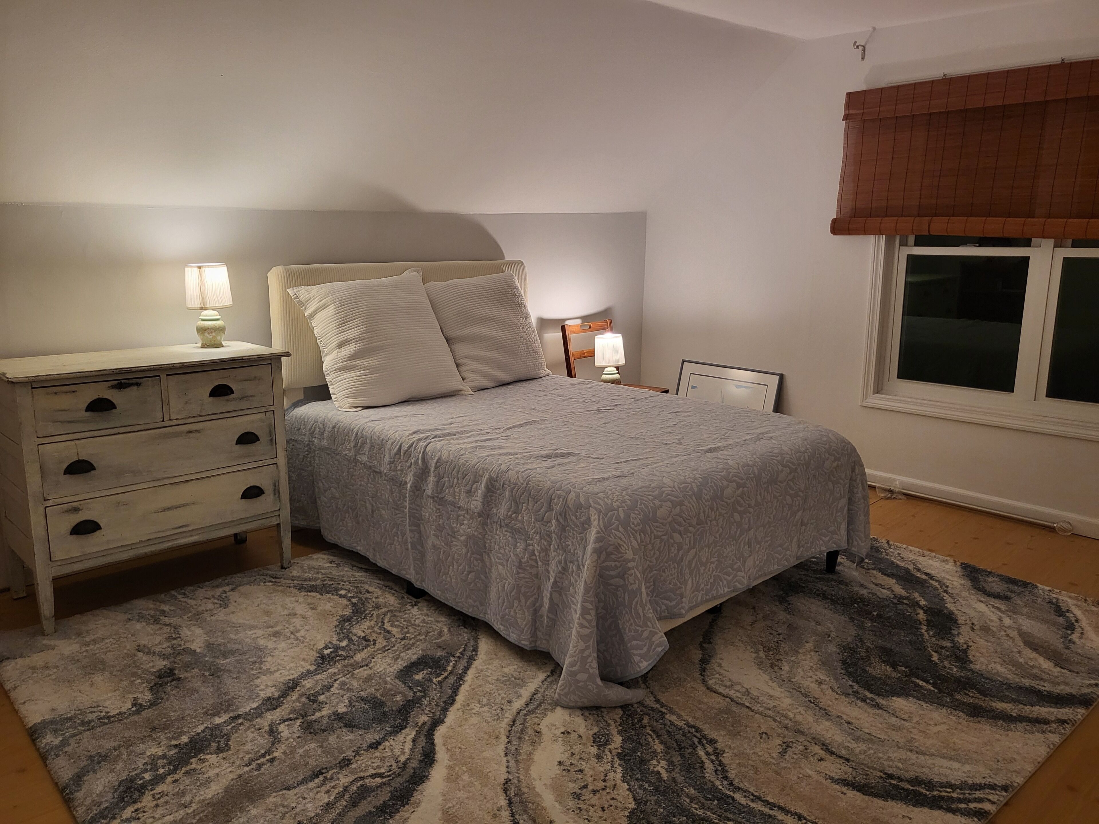 A bedroom with a bed, bedside tables, a nightstand, a window with a blind, and a patterned rug.