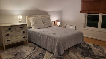 A bedroom with a bed, bedside tables, a nightstand, a window with a blind, and a patterned rug.