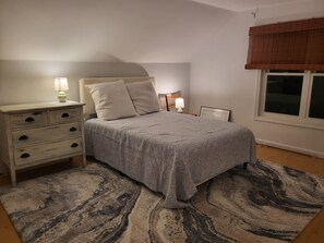 Ein Schlafzimmer mit Bett, Nachttischen, einem Fenster mit Jalousie und einem gemusterten Teppich.