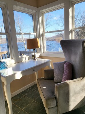 Ein Zimmer mit Schreibtisch, Stuhl und großen Fenstern mit Blick auf ein Gewässer.