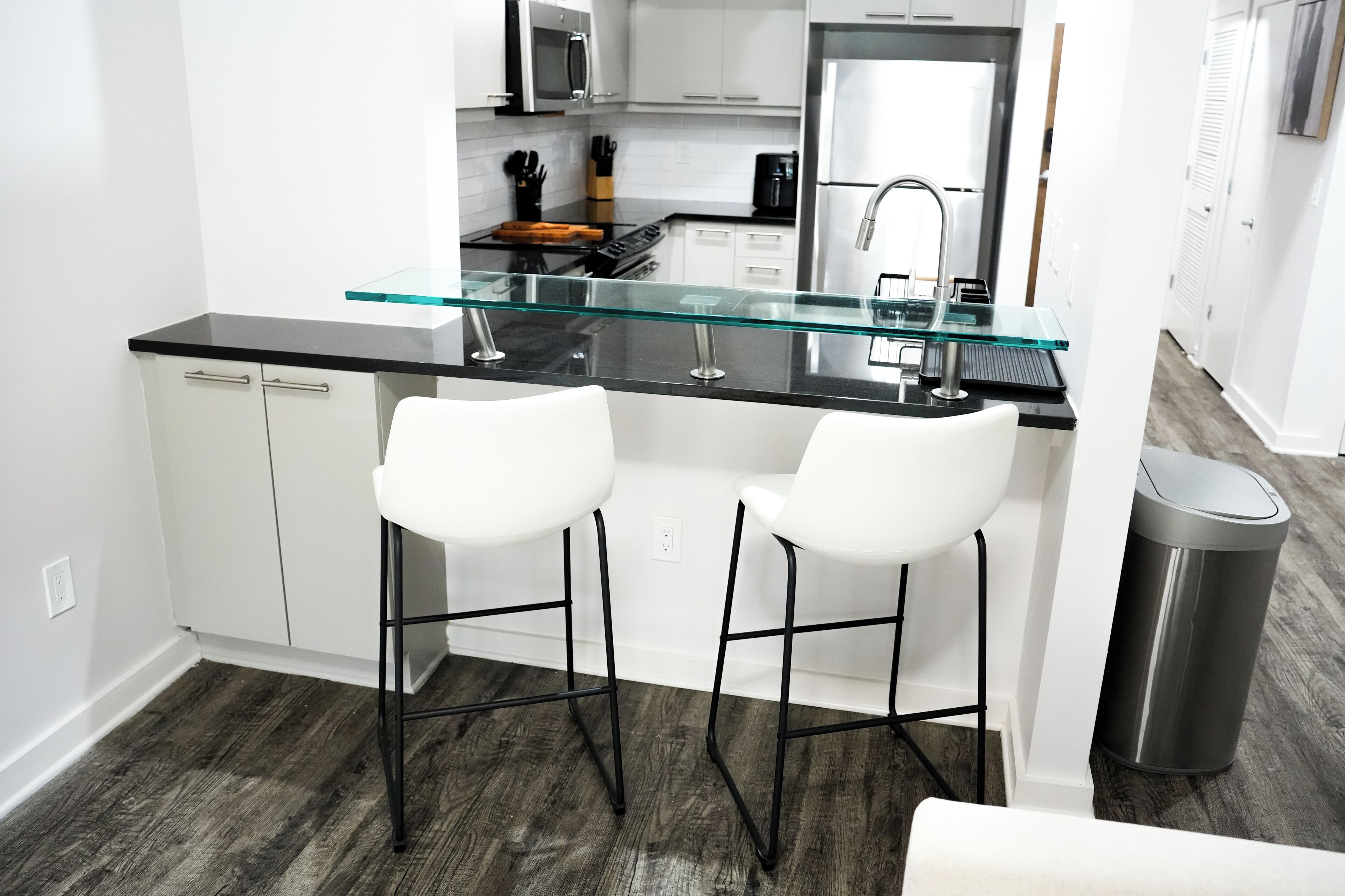 Una cocina moderna con una isla de vidrio, taburetes blancos de barra, electrodomésticos de acero inoxidable y un cubo de basura.