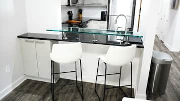 Una cocina moderna con una isla de vidrio, taburetes blancos de barra, electrodomésticos de acero inoxidable y un cubo de basura.