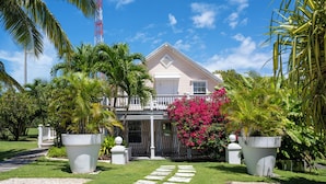 Una casa con fachada blanca, un balcón y un jardín cuidado con palmeras y una planta floreciente de color rosa.