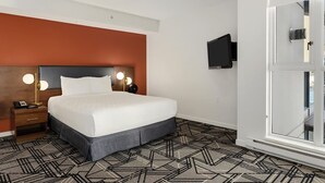 Habitación de hotel con cama, mesita de noche, televisor montado en la pared y vista a la ciudad a través de un ventanal amplio.