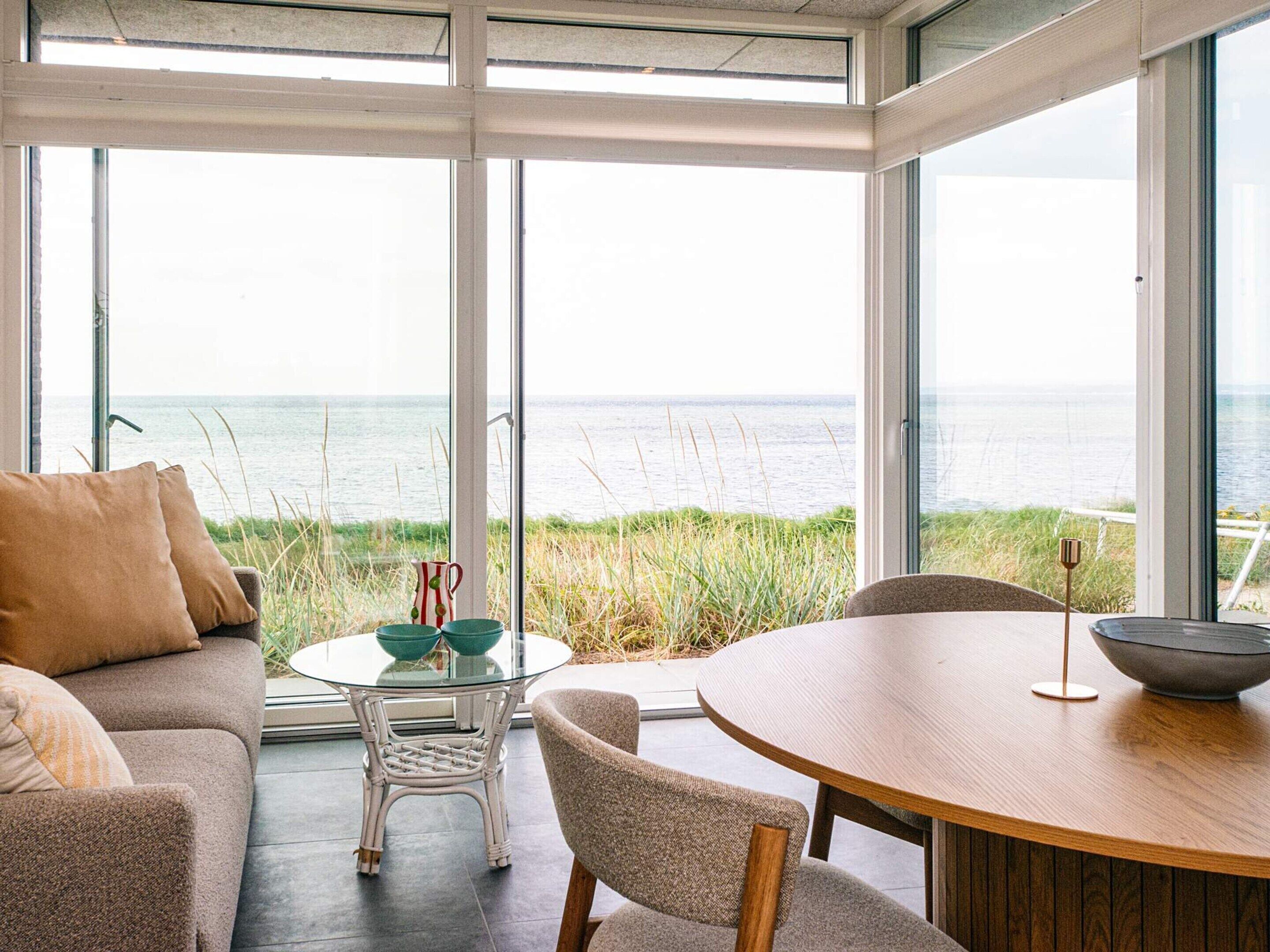 A modern interior with a glass table and chairs, a sofa, and a view of the sea through large windows.