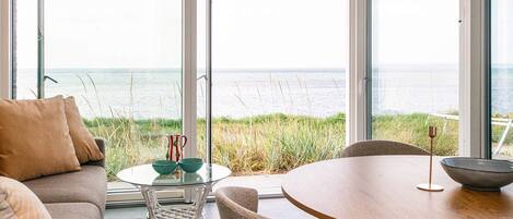 A modern interior with a glass table and chairs, a sofa, and a view of the sea through large windows.