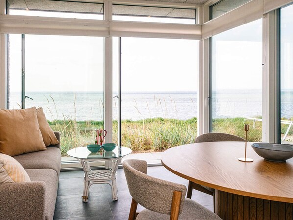 A modern interior with a glass table and chairs, a sofa, and a view of the sea through large windows.