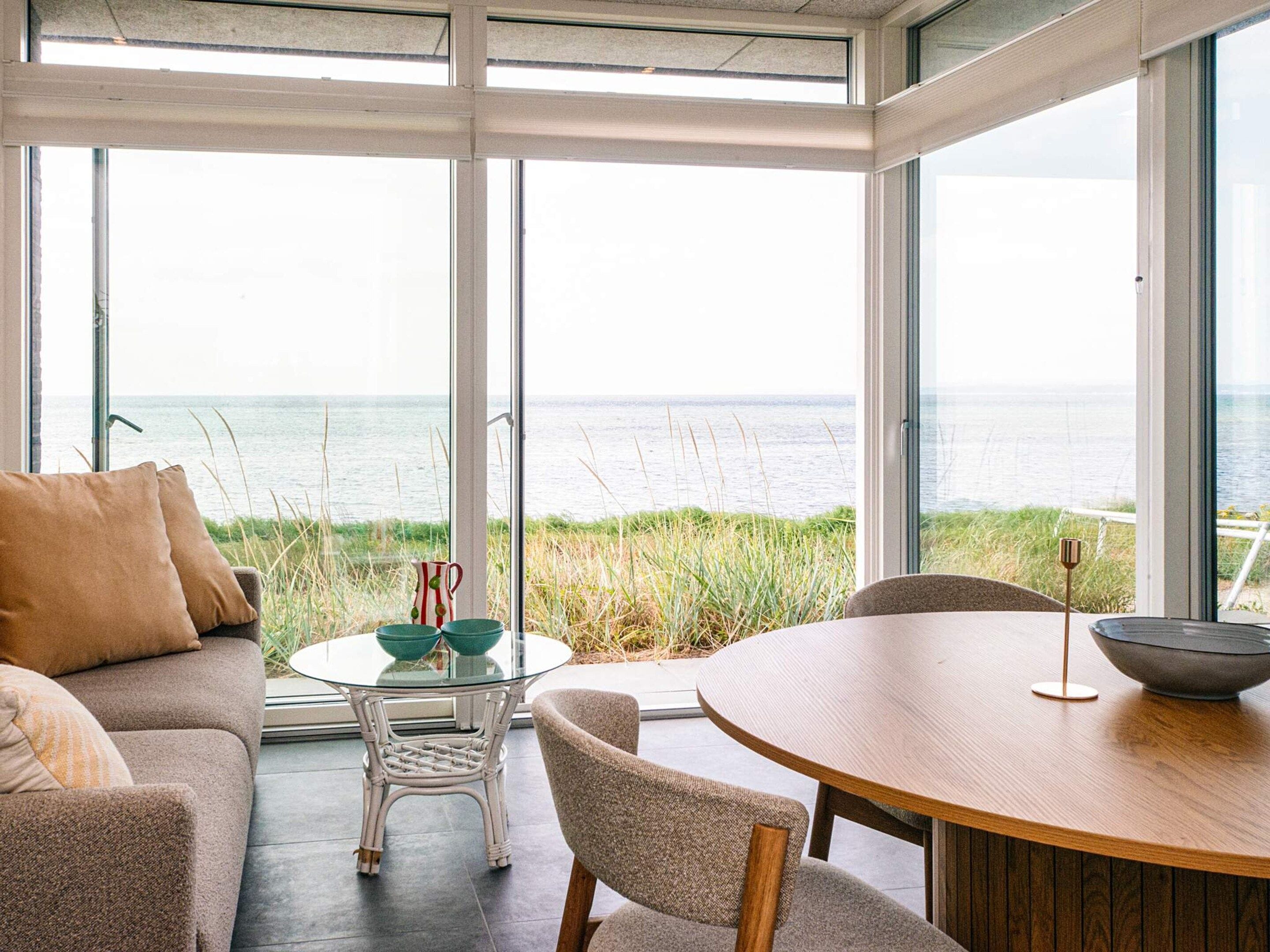 A modern interior with a glass table and chairs, a sofa, and a view of the sea through large windows.