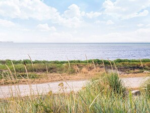 A coastal landscape with a sandy beach, grassy vegetation, and a calm sea under a partly cloudy sky.