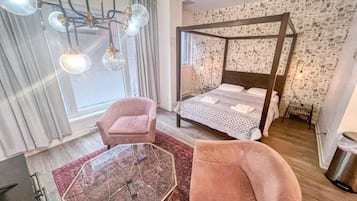 Una habitación de hotel moderna con una cama con dosel, una mesita de vidrio, dos sillones rosados y un ventanal con cortinas.