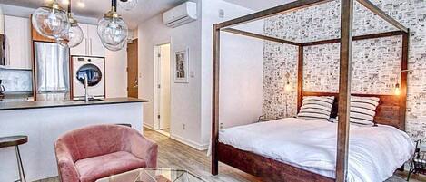 Un dormitorio moderno con cama con dosel, una mesita de centro de vidrio, un sillón rosa y una zona de cocina con barra.