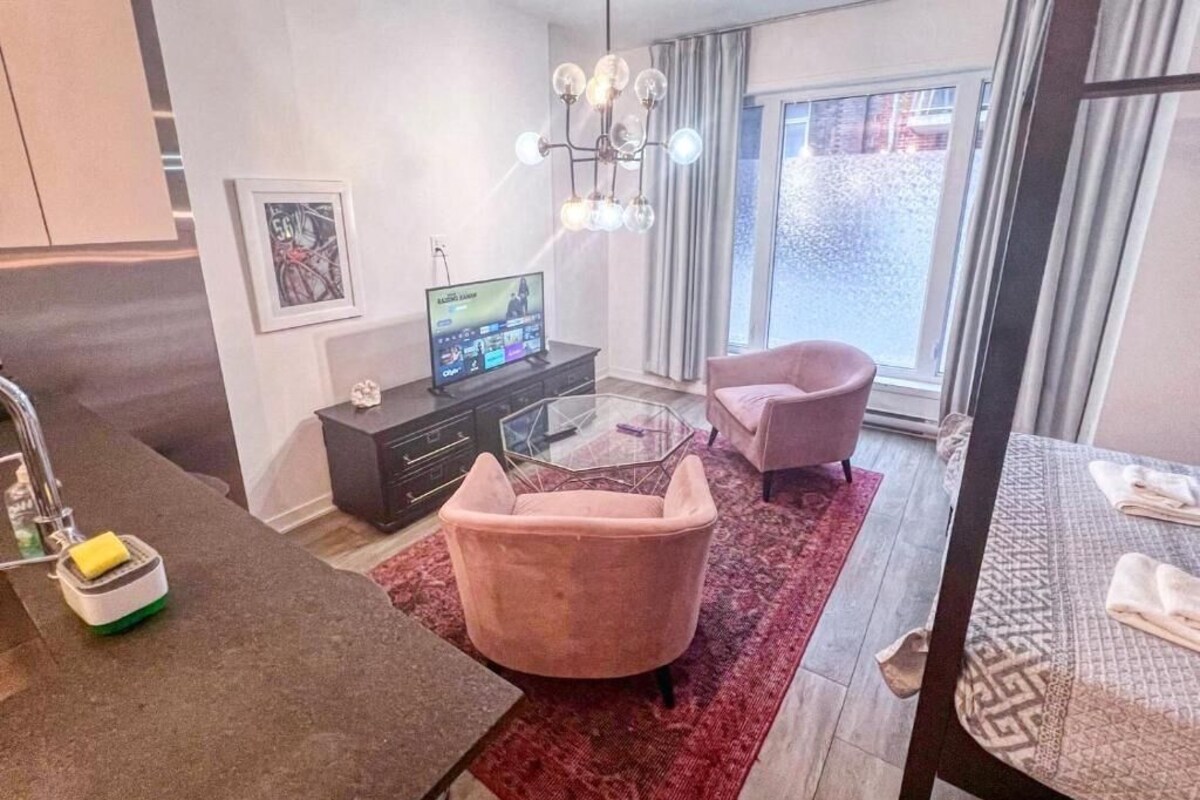 Una sala moderna con una mesa de centro de vidrio, un sillón rosa, un televisor y un ventanal con cortinas.
