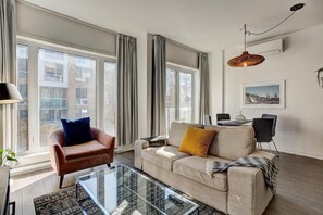 A modern living room with a sofa, armchair, glass coffee table, and a dining area with chairs. - The Downtown Zando (Montréal)