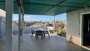 Een overdekt terras met een tafel en stoelen, uitzicht op een heuvel en een gebouw ingang.