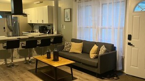 A living room with a sofa, coffee table, and chairs, a kitchen in the background, and a door with a window. - Luxury Stay Gastonia (Gastonia)