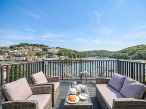 Un balcón con mobiliario de mimbre, una mesa con alimentos para el desayuno y una vista panorámica de un pueblo y colinas.