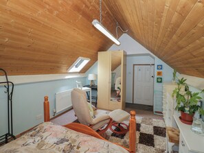Um quarto com teto inclinado, uma cama com colcha florida, uma cadeira, uma escrivaninha com abajur, um radiador e uma porta com uma planta.