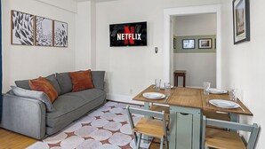 Olohuoneessa on harmaa sohva, puinen ruokapöytä tuoleineen, taulutelevisio ja seinällä on kehystettyjä kuvia.