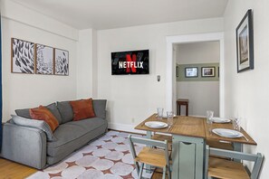 Ein Wohnzimmer mit grauem Sofa, einem Holzesstisch mit Stühlen, einem Flachbildfernseher und gerahmten Bildern an der Wand.