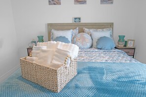 A neatly made bed with a blue patterned bedspread, a wicker basket with folded towels, and decorative pillows with sea-themed designs. - Lehigh Fun Home (Lehigh Acres)