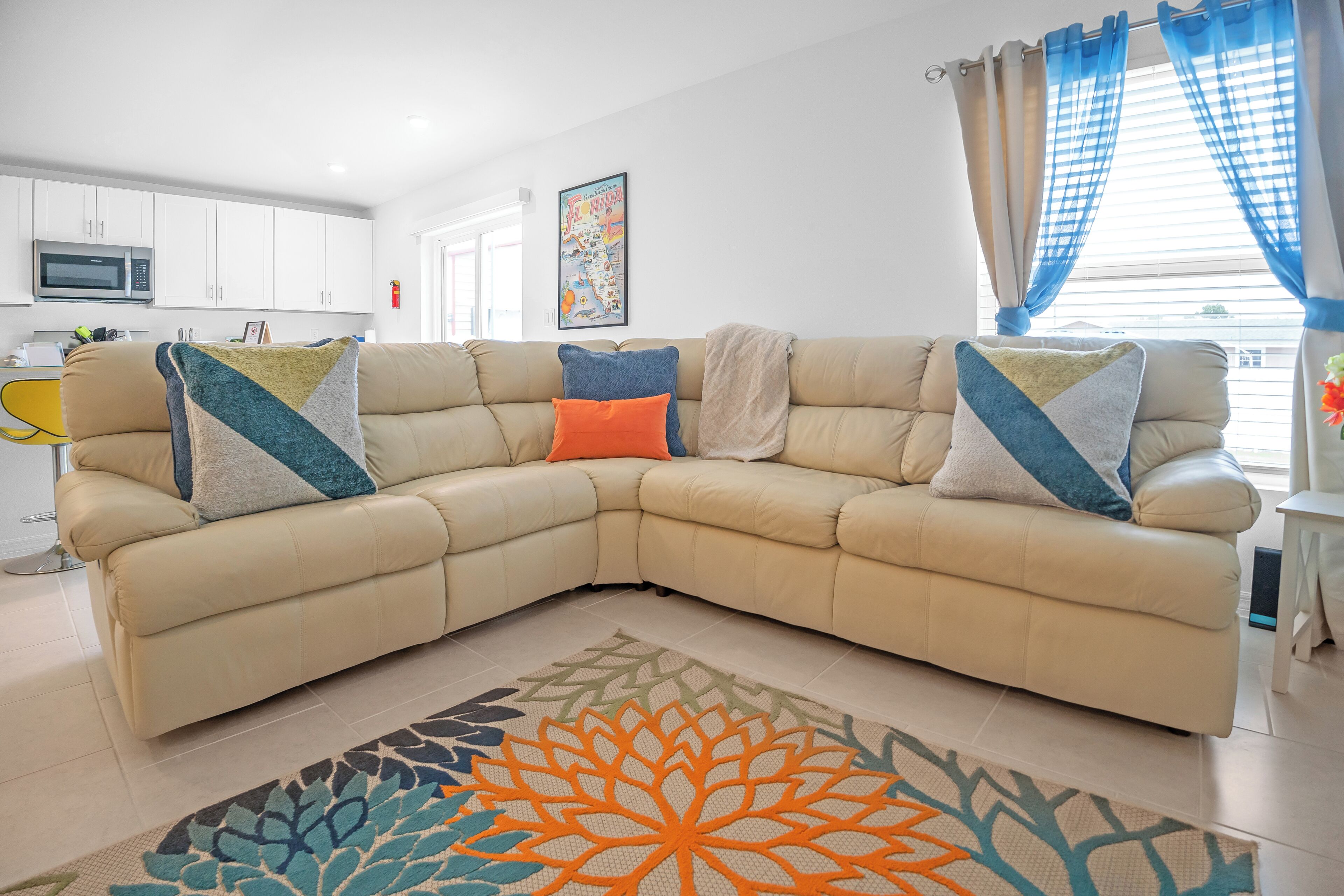 Ein Wohnzimmer mit einem beigefarbenen Ecksofa, einem gemusterten Teppich, einer Küche im Hintergrund und einer gerahmten Karte an der Wand.