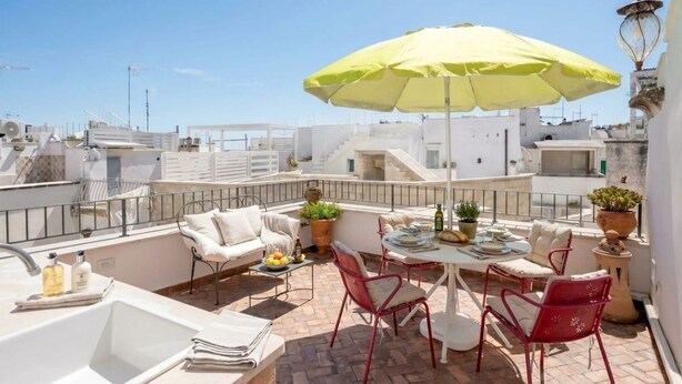 Una terraza en la azotea con una sombrilla amarilla, muebles blancos, un lavamanos y plantas en macetas.