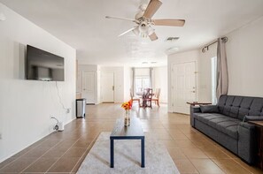 A modern living room with a sofa, a coffee table, a TV mounted on the wall, a ceiling fan, and a dining area in the background. - 3-bedroom house in fabulous Tucson with cool AC for relaxing stay (Tucson)