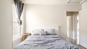 Een slaapkamer met een bed, een nachtkastje, een raam met gordijnen en een deur naar een andere kamer.