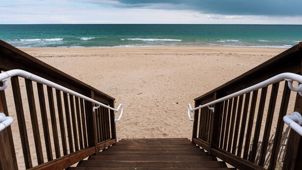 Un escalier en bois mène à une plage de sable, d’où l’on peut admirer l’océan et le ciel nuageux.