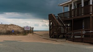 Un bâtiment situé au bord de la plage, avec un escalier menant à un balcon, une pancarte indiquant que la plage est fermée, et une zone de plage sablonneuse.