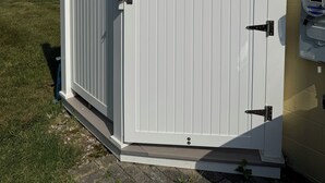 Une porte en métal blanc, dotée d’un loquet, est fixée à un bâtiment dont la façade est de couleur beige.