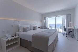 A hotel room with a bed, desk, chair, and a view of the ocean. - Elegant Beachfront Suite Perfect for a Romantic Getaway in Rio! (N/A)