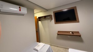 Ein Hotelzimmer mit Bett, einem an der Wand befestigten Fernseher, einem Holzregal und einer Klimaanlage.