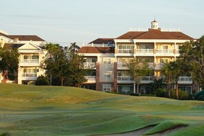 Ein mehrstöckiges Hotel mit Balkonen, vor einer hügeligen Landschaft und einem Golfplatz im Vordergrund.