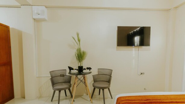 A hotel room with a bed, a TV mounted on the wall, two chairs around a small table with a plant, and a wooden door.