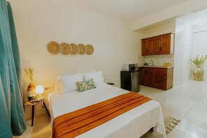 A hotel room with a bed, a bedside table with a lamp, a small plant, and a kitchen area with wooden cabinets and a sink.