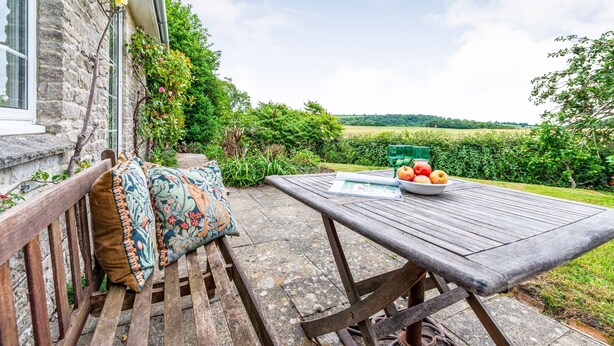A wooden bench with cushions, a table with a bowl of fruit, and a view of a green field and trees. - Greygles (Sleeps 5) -Melcombe Bingham, Dorchester sleeps 5 guests  in 3 bedrooms (Melcombe Bingham)