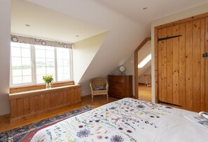 Ein Schlafzimmer mit großem Fenster, einem Bett mit floralem Überwurf, einer Holzkommode und einem Sessel.