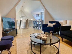 Ein modernes Wohnzimmer mit Fernseher, Sitzgelegenheiten und einem Essbereich im Hintergrund.
