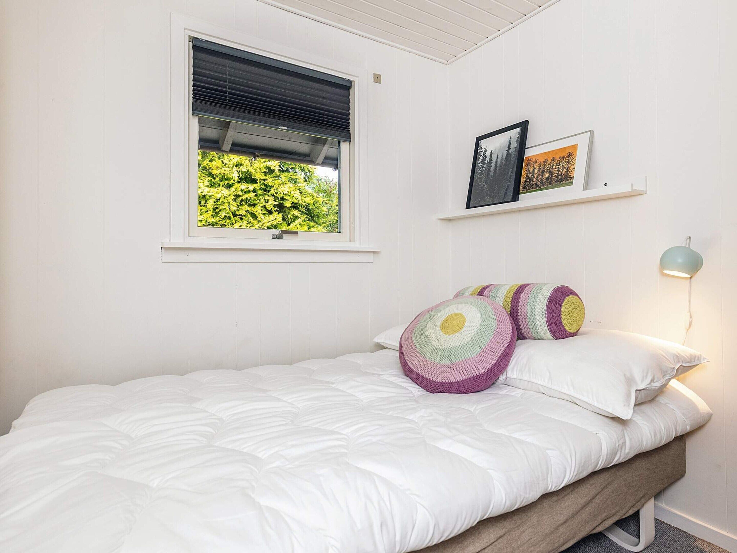 Quarto com cama, janela com persianas, prateleira na parede com fotos e abajur.