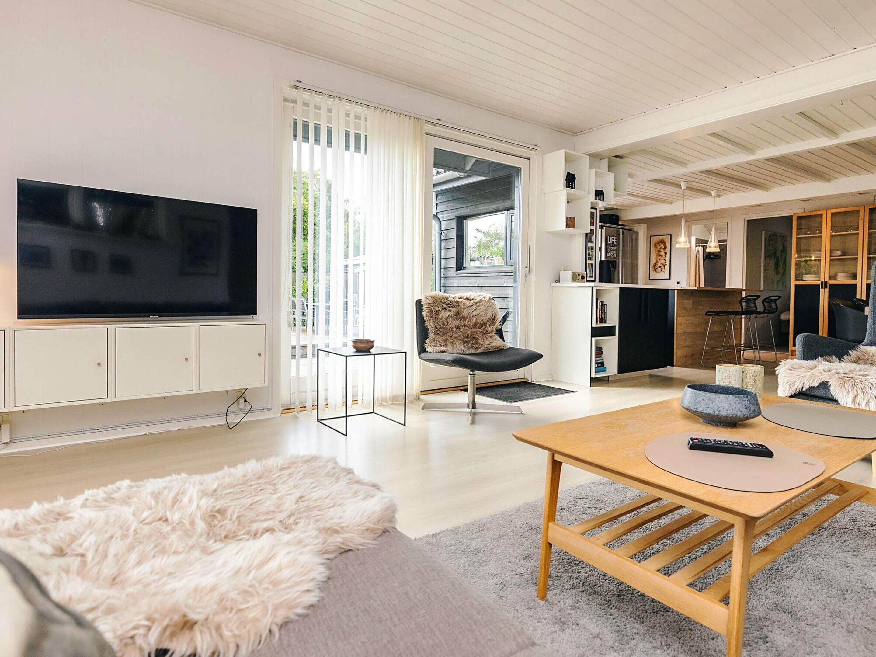 Uma sala de estar moderna com uma TV de tela plana, uma mesa de centro de madeira, um tapete branco felpudo e um espaço de estar confortável.