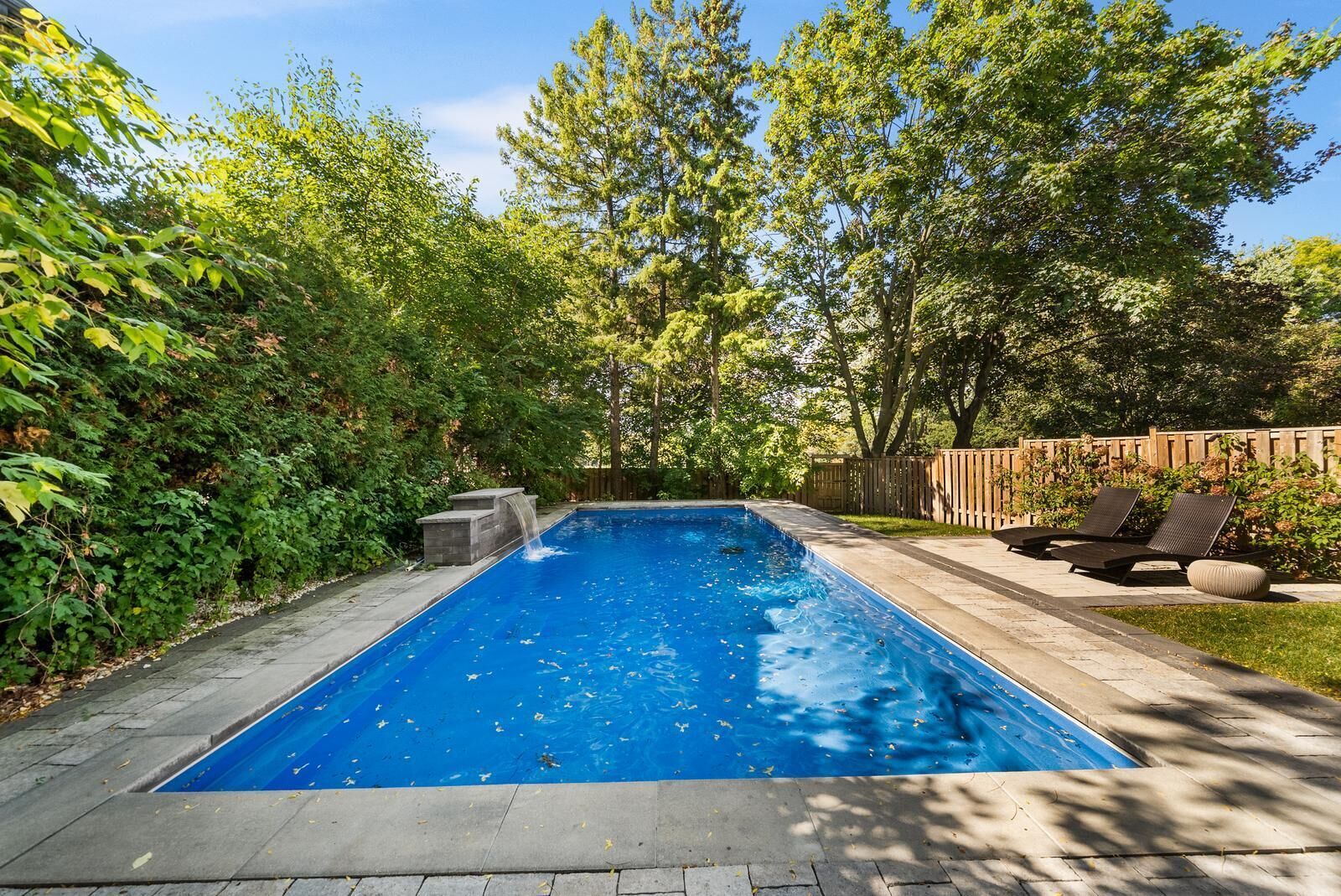 Una piscina rettangolare con cascata, circondata da vegetazione lussureggiante e un pontile in legno con mobili da esterno.