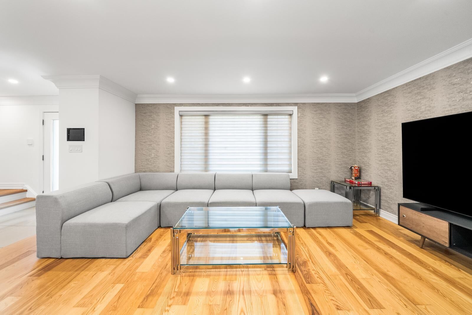 A modern living room with a sectional sofa, a glass coffee table, a flat-screen TV, and wooden flooring.