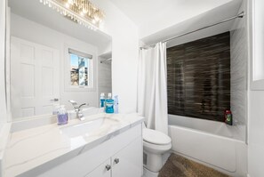 Ein Badezimmer mit weißem Waschtisch, Toilette, Duschkabine mit Vorhang und einem Fenster mit Jalousien.