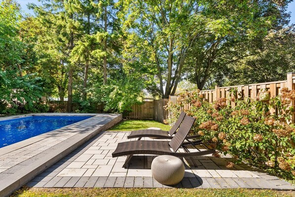 A swimming pool with lounge chairs and a round ottoman, surrounded by trees and a wooden fence.