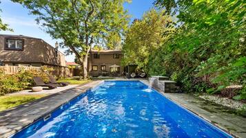 A rectangular swimming pool with a waterfall feature, surrounded by a wooden deck and lounge chairs.